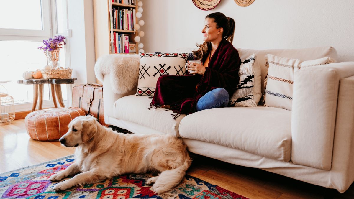 A woman sits on a sofa sipping a drink while her dog sits calmly on a rug by her feet.